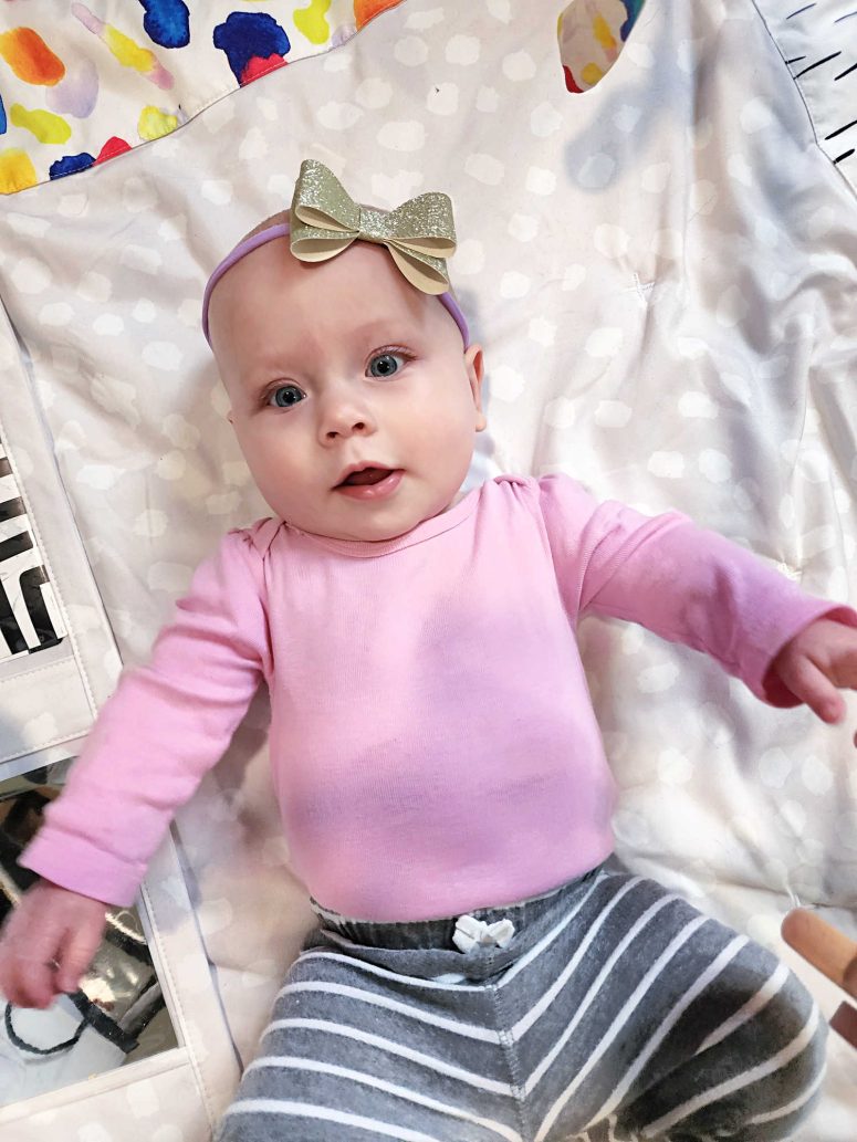 baby girl in a pink shirt and grey striped pants wearing a gold glitter bow