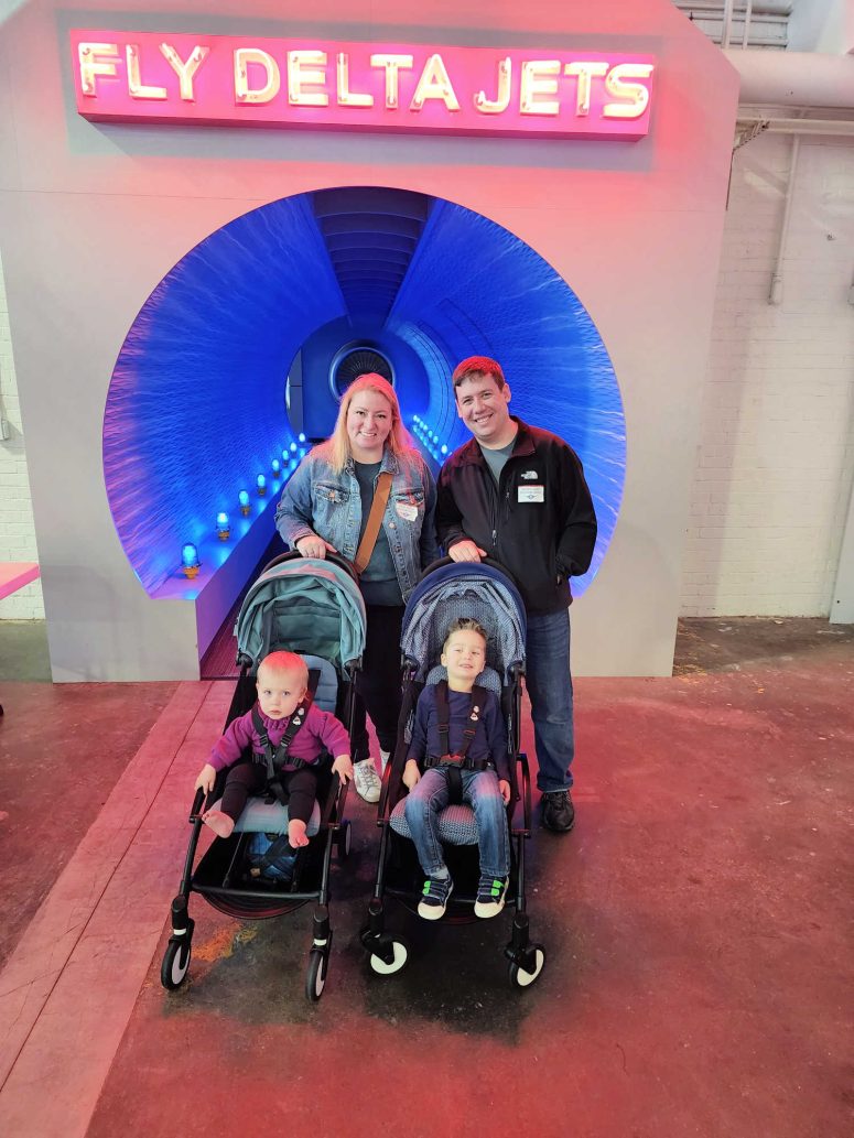 A mom and dad with their toddlers standing in front of a blue tunnel with "Fly Delta Jets" illuminated in Red above