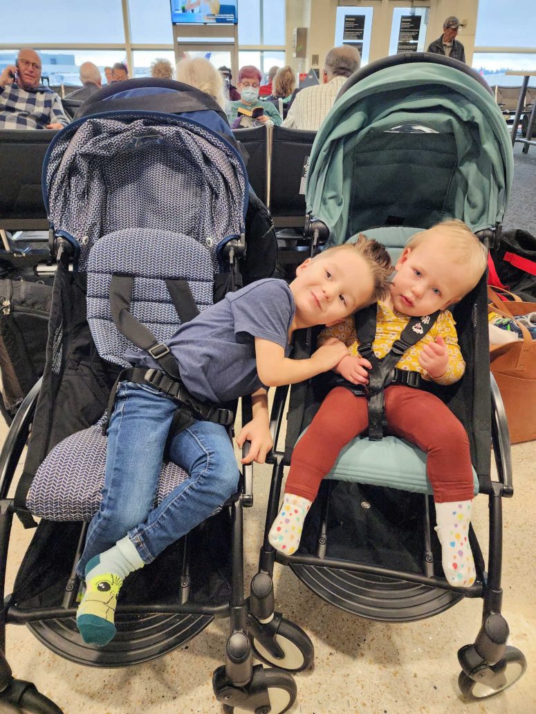 A toddler boy hugging his younger sister while they are seated in separate strollers.