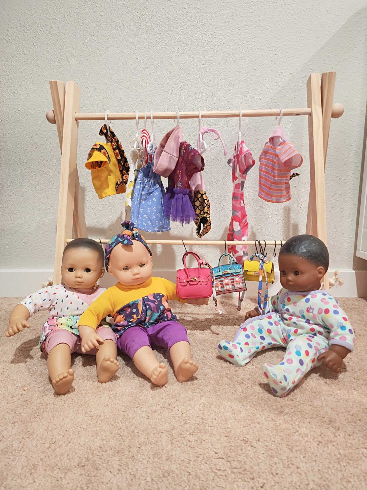 Three dolls sit in front of a triangle shaped wooden structure with clothes and purses hanging.