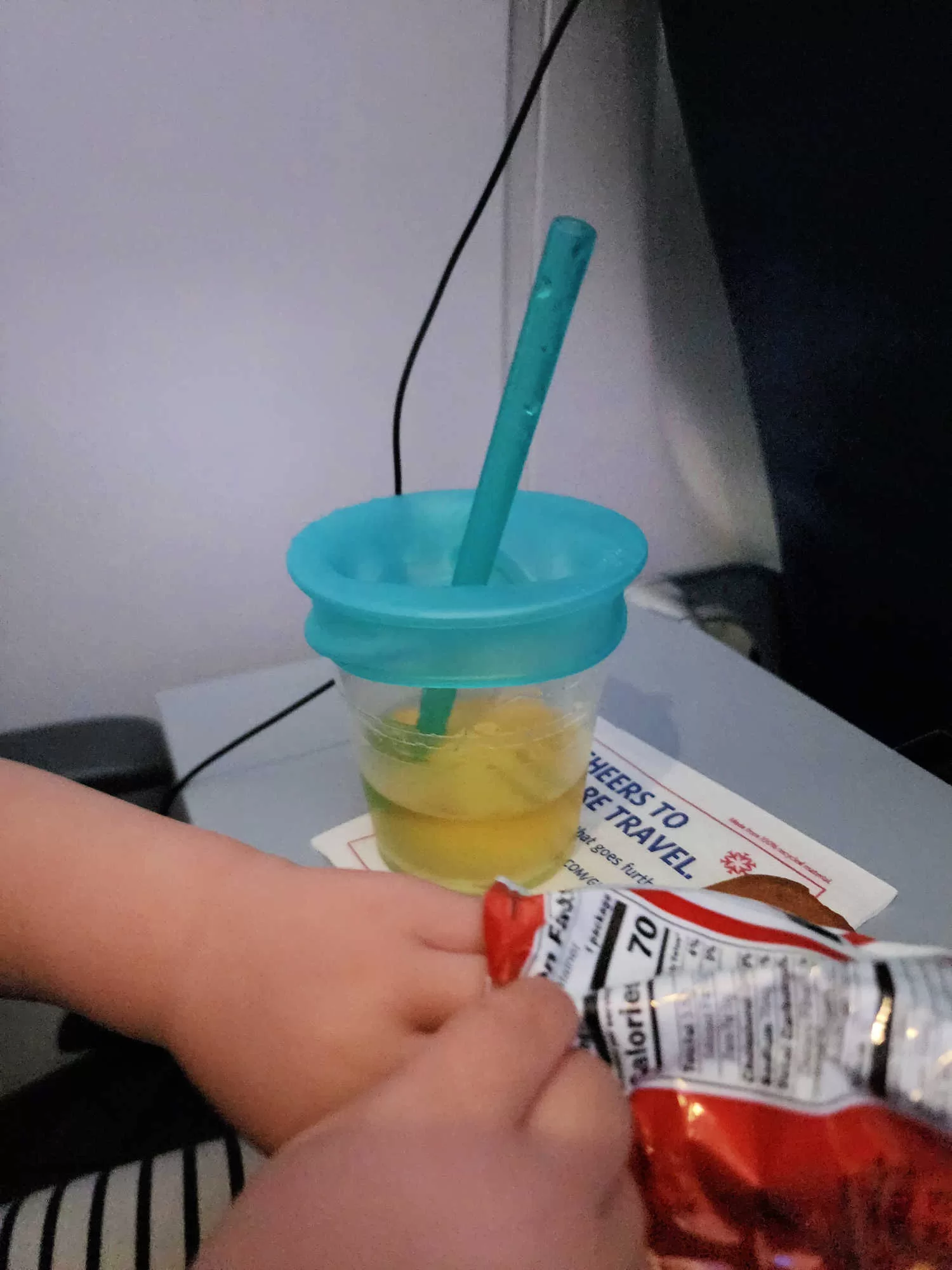 A cup of apple juice sits on the airplane tray table. Over the cup is a silicone cover with straw. 