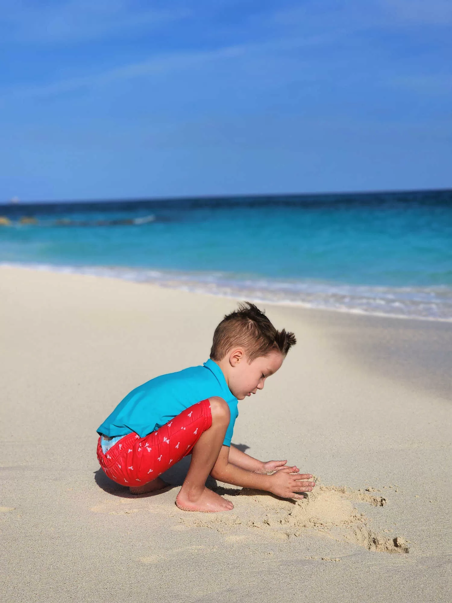 A four year old boy plays in the sand in the Bahamas. He is wearing red swim suit with a blue rash guard.