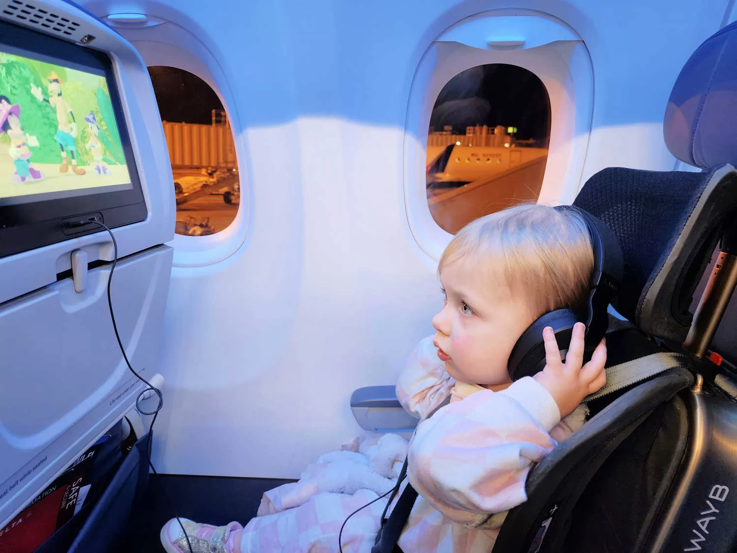 A two year old girl sits in a car seat on the airplane. She has earphones on and is watching Mickey Mouse on the tv. 