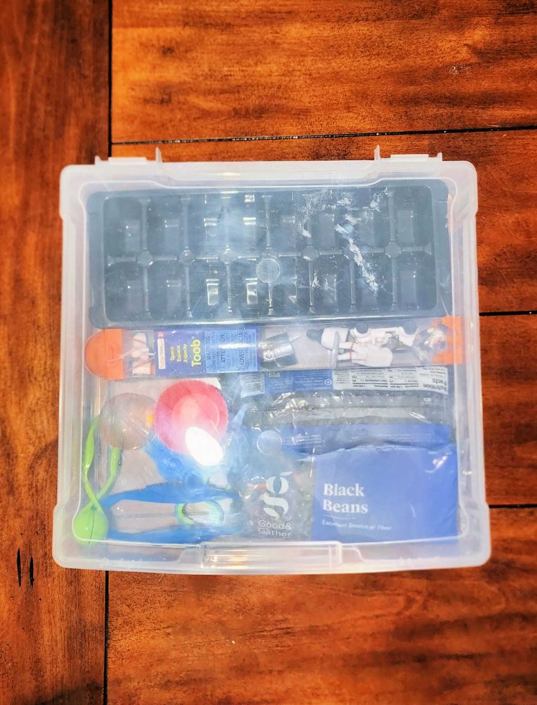 A white 12x12 box with sensory bin items including an ice cube tray, black beans, space toys, cupcake liners, and various tweezers.
