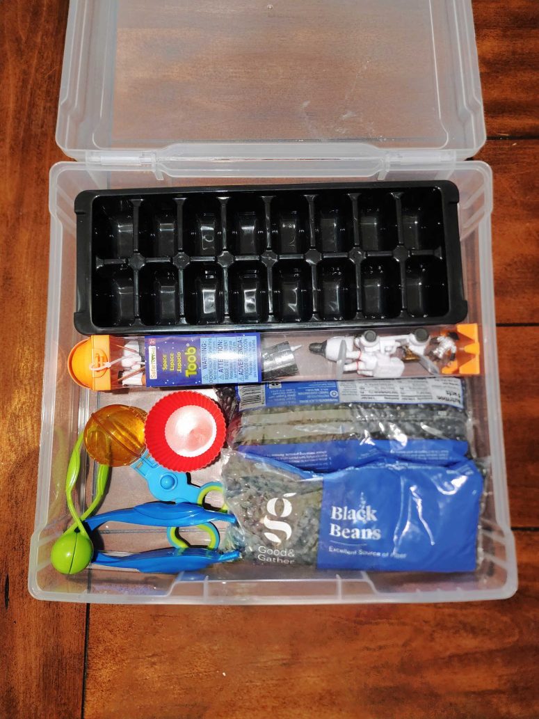 A white 12x12 box with sensory bin items including an ice cube tray, black beans, space toys, cupcake liners, and various tweezers.